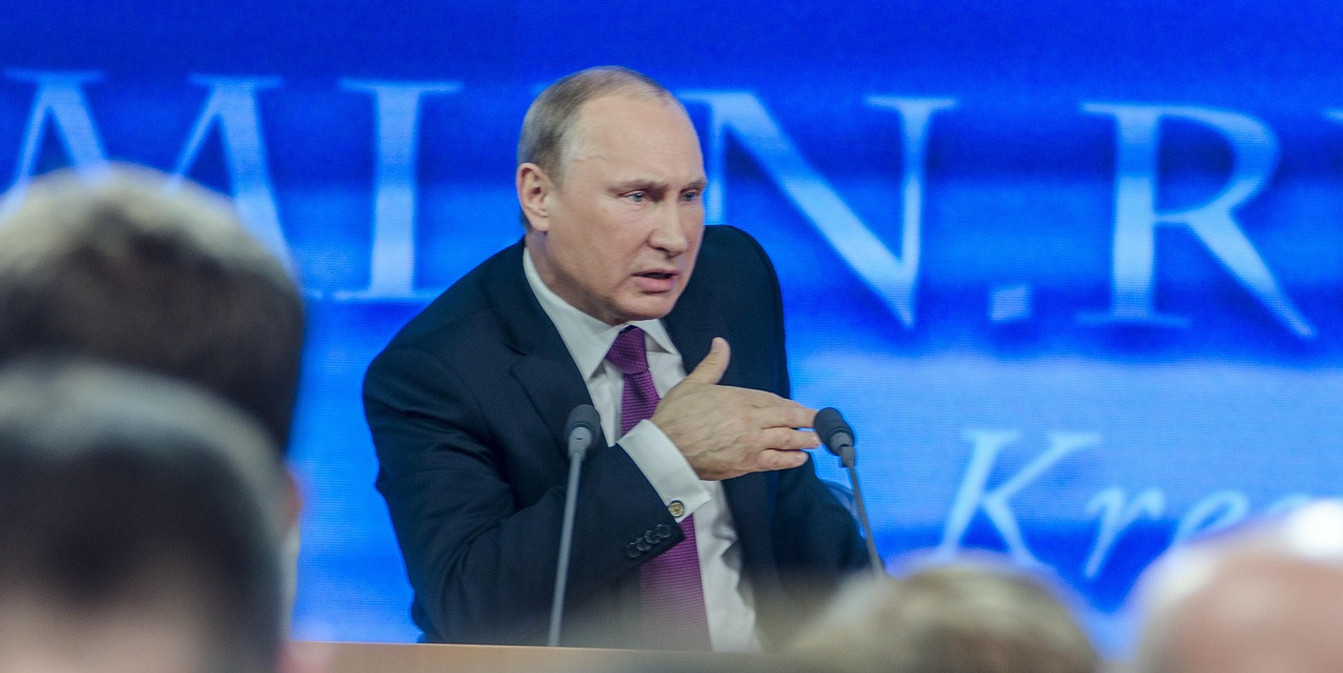 Владимир Путин выступает за трибуной на фоне синего экрана с надписью Kremlin. Президент России в темном костюме и галстуке активно жестикулирует во время речи.