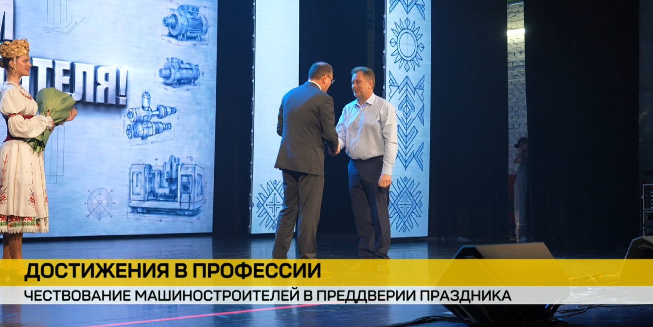 Чествование машиностроителей: двое мужчин пожимают руки на сцене на фоне экрана с чертежами и орнаментом. Слева стоит девушка в белорусском национальном костюме с букетом.