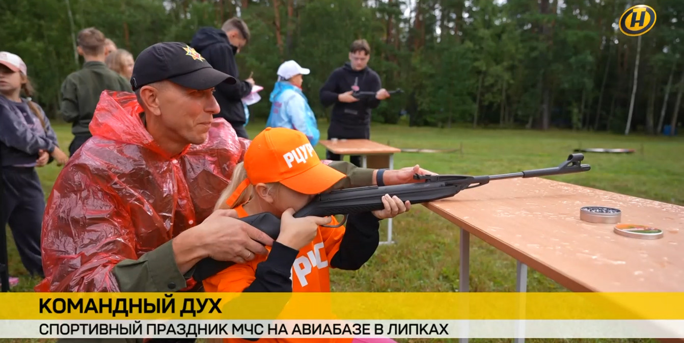 Взрослый мужчина в красном дождевике помогает девочке в оранжевой кепке целиться из пневматической винтовки. На заднем плане — лесной пейзаж и другие участники праздника МЧС.