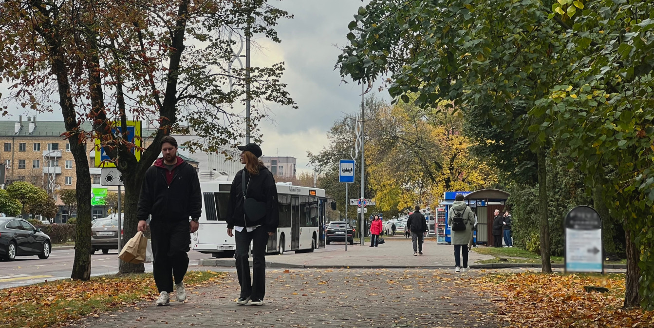 Люди гуляют по усыпанному желтыми листьями тротуару вдоль городской улицы, на заднем плане виден белый автобус.
