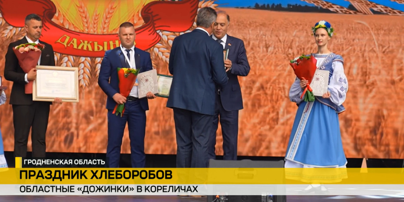 Областные «Дожинки» в Кореличах: на сцене Гродненской области награждают передовиков-хлеборобов. Мужчины в костюмах с грамотами и цветами, рядом девушка в белорусском народном наряде.
