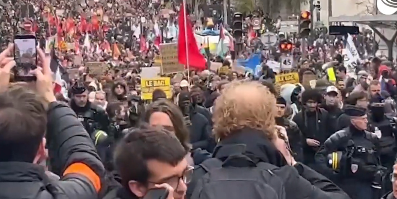 Огромная толпа протестующих на улице Парижа с плакатами и флагами. На переднем плане люди снимают происходящее на мобильные телефоны, видны сотрудники полиции в экипировке.