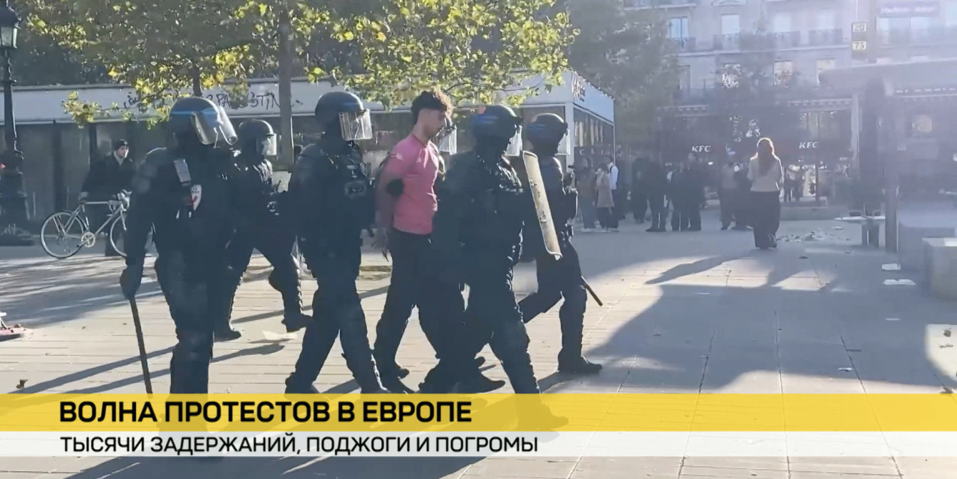 Полицейские в защитной экипировке задерживают мужчину в розовой футболке во время протестов в Европе. На фоне видна улица и вывеска KFC.
