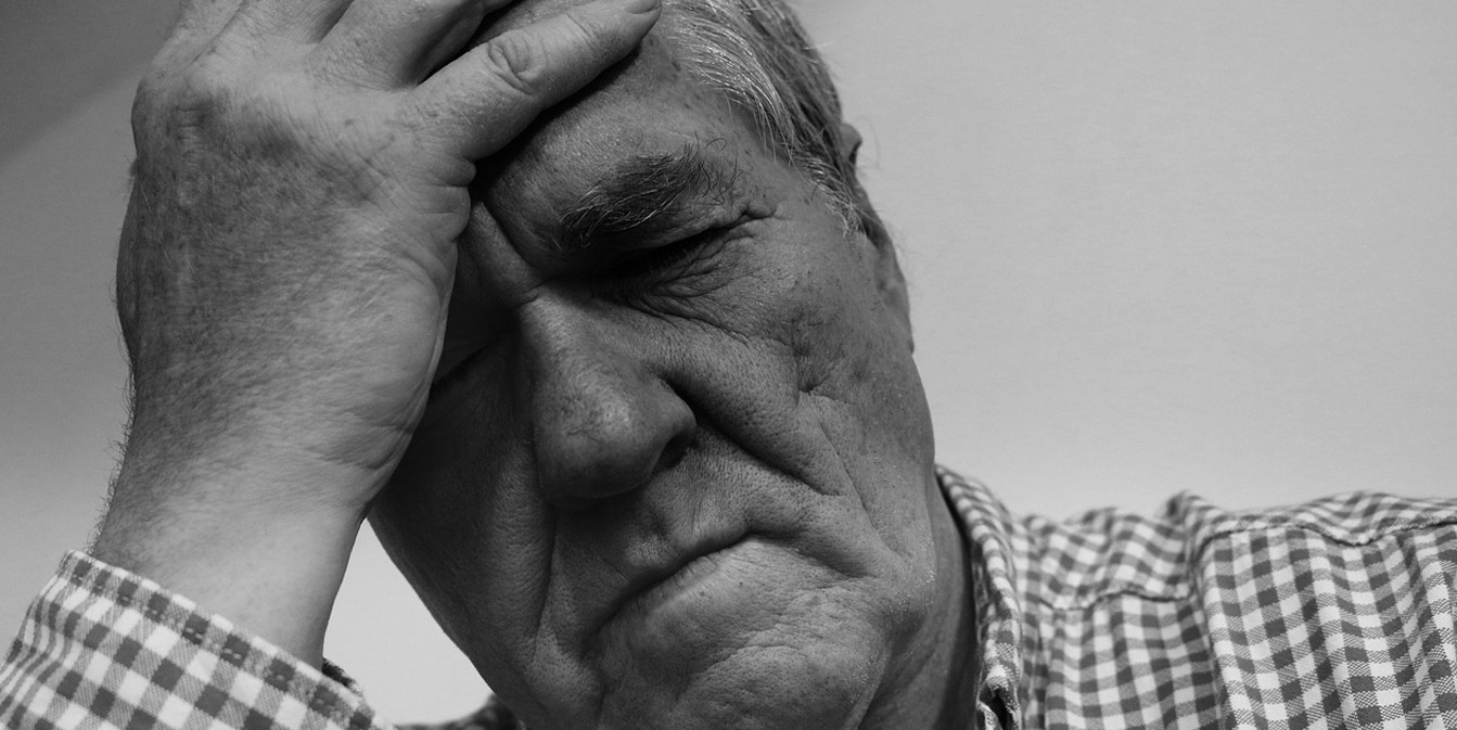 Черно-белое фото крупным планом: пожилой мужчина в клетчатой рубашке зажмурился и прижал руку ко лбу, испытывая сильную головную боль.