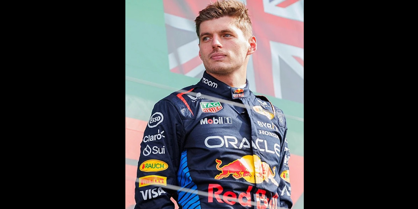Макс Ферстаппен в темно-синем гоночном комбинезоне Red Bull Racing стоит на подиуме и смотрит в сторону. На заднем плане виден фрагмент флага Великобритании.