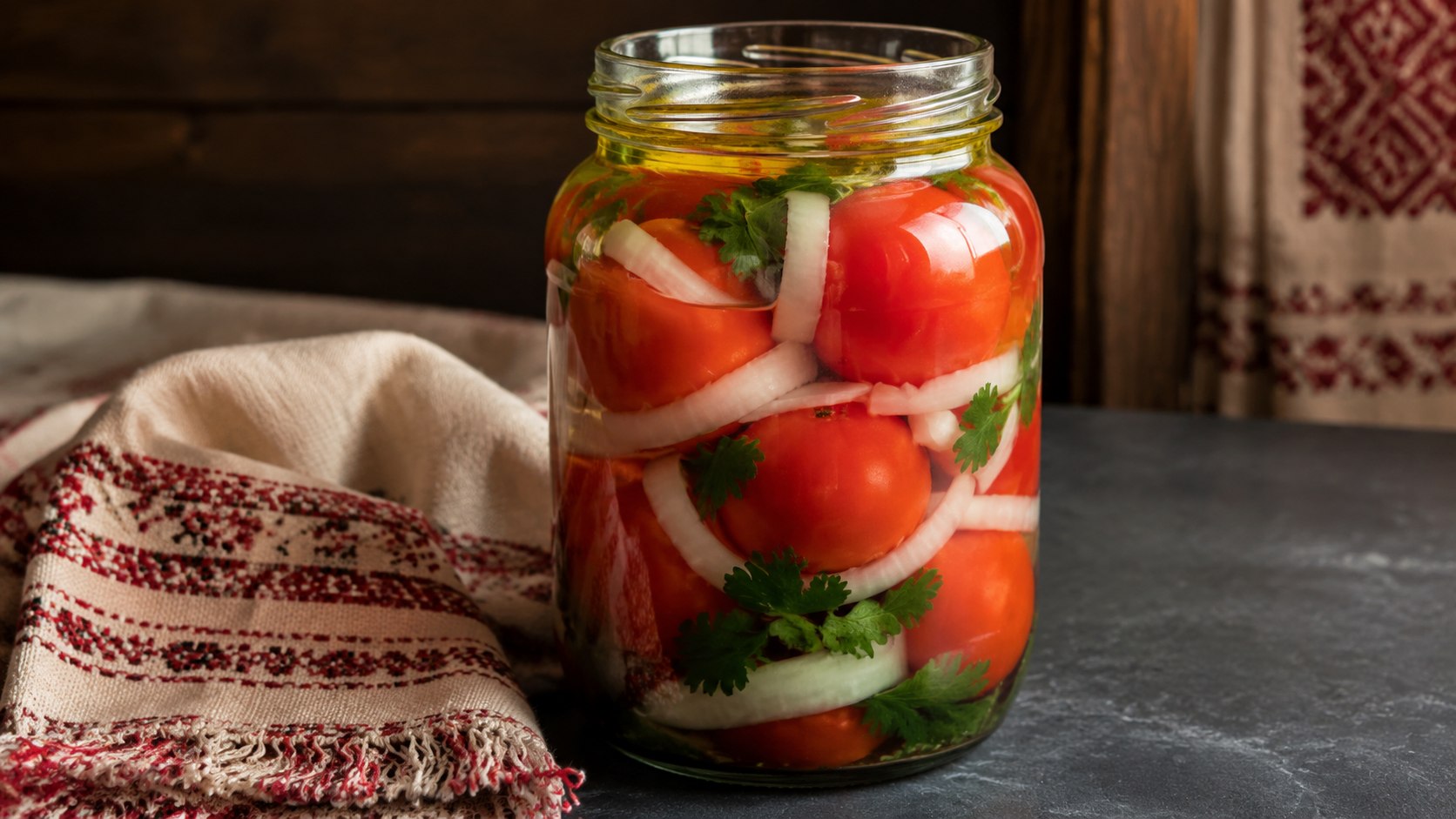 A hyper-realistic close-up shot of a 1-liter glass jar filled with marinated red tomatoes. The tomatoes are ripe, plump, and intact, neatly layered with thick white onion rings and fresh green parsley sprigs. A thin, glistening layer of vegetable oil is visible at the top of the clear liquid. The jar is placed on a modern dark slate countertop next to a vintage белорусский linen napkin with red embroidery patterns. Beautiful warm side lighting accentuates the glossy texture of the glass and the rich red color of the tomatoes. Professional food photography, 8k, sharp focus, highly detailed, vivid colors, editorial style.