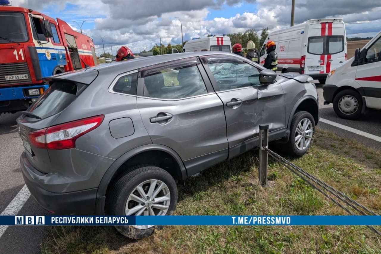 серый кроссовер Nissan Qashqai, врезавшийся в металлическое тросовое ограждение на разделительной полосе дороги. На месте аварии работают спасатели МЧС у пожарной машины и медики возле автомобилей скорой помощи. Внизу снимка расположена плашка МВД Республики Беларусь.