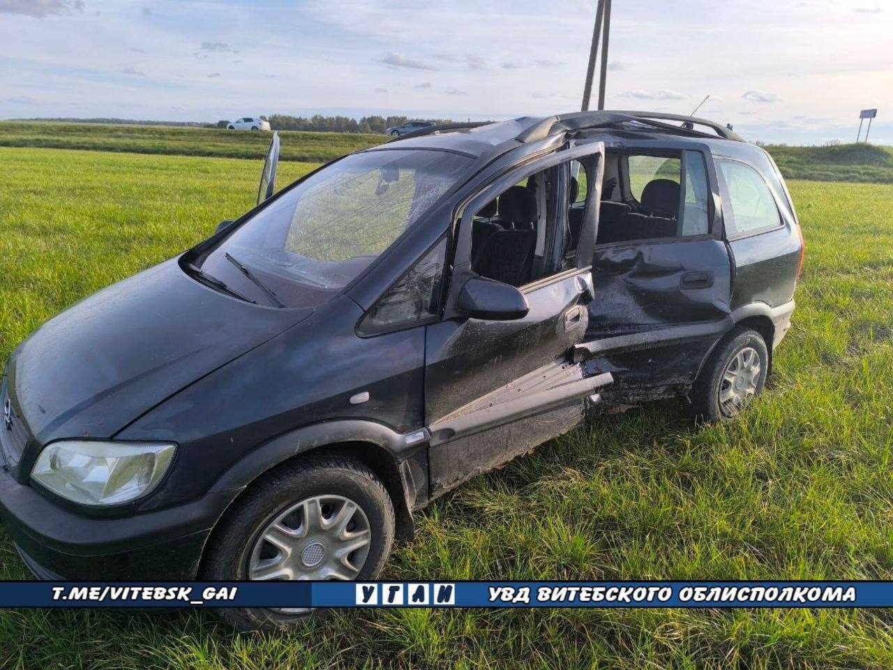Черный минивэн Opel Zafira со значительными повреждениями левого борта стоит в зеленой траве в кювете у дороги. Вся водительская сторона сильно деформирована, передняя дверь выгнута наружу, боковые стекла выбиты, а крыша в районе центральной стойки смята.